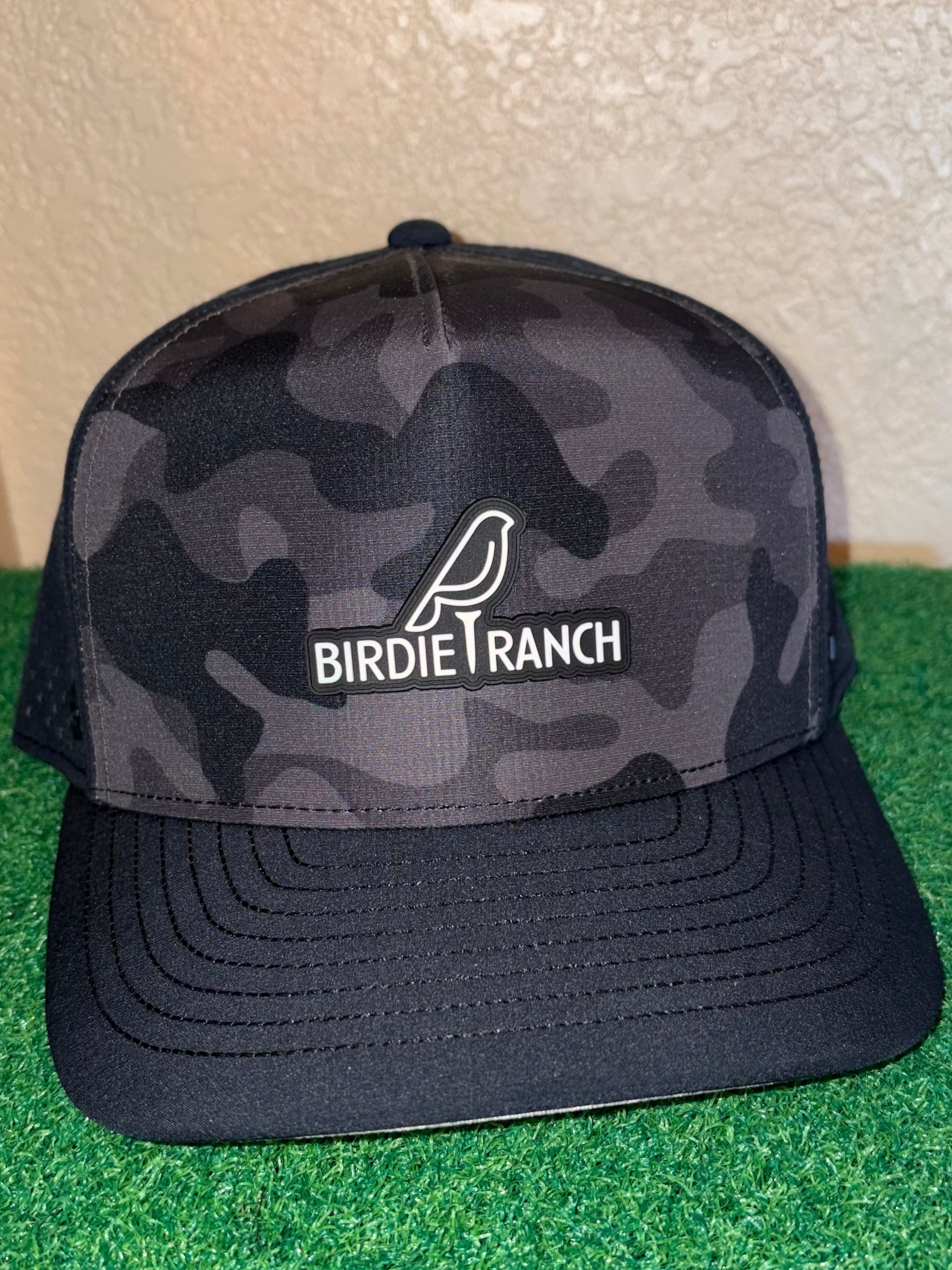 Black camouflage cap with 'Birdie Ranch' logo on a green surface
